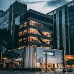 Night view of a city block with illuminated store, modern architecture, and cloudy sky.